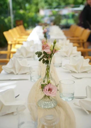 Esküvői asztaldísz rózsaszín és fehér virágokkal, elegánsan terített kültéri asztalon.